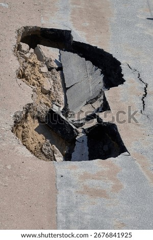 Large sinkhole in the ground exposing different layers of earth and concrete, revealing a damaged street surface in a construction area.