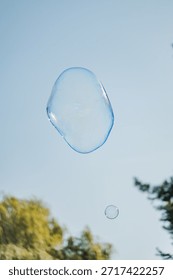 A large and a small soap bubble float in the air in front of a blue sky. In the background you can see some trees.