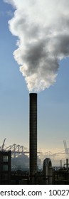 Large Smoke Stack Spewing Steam With Shipping Port Cranes In The Background And Blue Sky Above./ Port Of Seattle Smoke Stack /  Industrial Output.