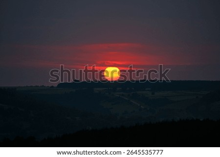 A large solar disk dips below the horizon, painting the sky in hues of orange and purple. Abstract designs. Gorgeous hot summer sunset with big red sun circle. Photo wallpaper. Beauty of earth.