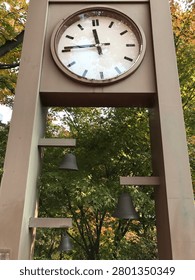 A large square clock faces forward on a metal structure adorned with three hanging bronze bells against green and autumn trees.