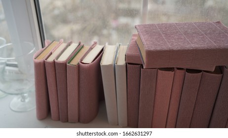 Large stack of books on windowsill. Books wrapped in colored paper lie on window sill.