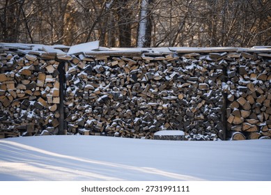 Large stack of cut firewood covered with snow during winter. Outdoor wood storage in a rural setting with wooden logs neatly piled and snow-covered ground in cold weather.