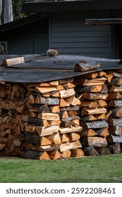 Large stack of freshly cut and split logs into firewood, covered to protect from weather, on lawn in side yard of house, winter heat source
