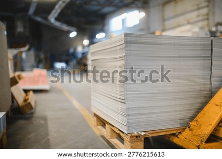Large stack of raw paper sheets resting on a wooden pallet in a busy publishing and industrial workshop