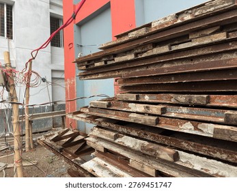 A large stack of rusty, weathered metal and wood planks sits on a dirt ground next to a building with red and grey walls, surrounded by tangled electrical wires.