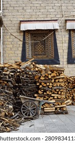 A large stack of split firewood logs with an old wooden wheelbarrow parked in front, set against a traditional Tibetan brick wall with an ornately decorated window at a temple in Shangri-La, Diqing Tibetan Autonomous Prefecture, Yunnan, China.(Oct,12,2025)