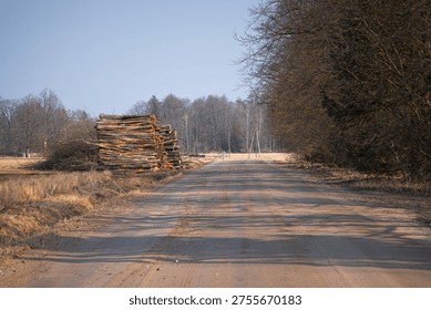 Large stack of timber logs piled by dirt road in rural spring landscape
