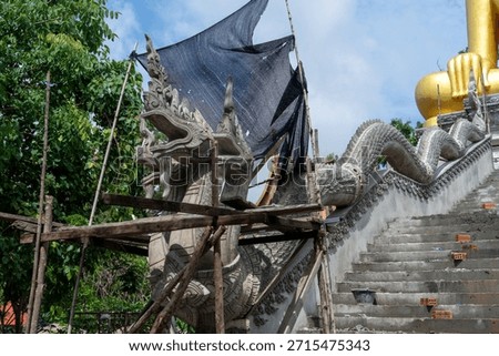 A large statue under construction features dragon sculpture and scaffolding, surrounded by greenery and blue sky. scene captures blend of artistry and craftsmanship in serene environment