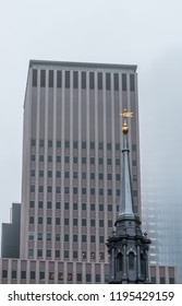 A Large Steeple With A Gold Bird Weathervane Stands In Front Of A Taller Modern Building. Low Clouds And Fog Are Part Way Down The Face Of The Modern Building.