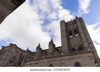 A large stone building with a clock tower and a steeple. The building is surrounded by a stone wall and has a large archway. The sky is cloudy and the sun is shining through the clouds