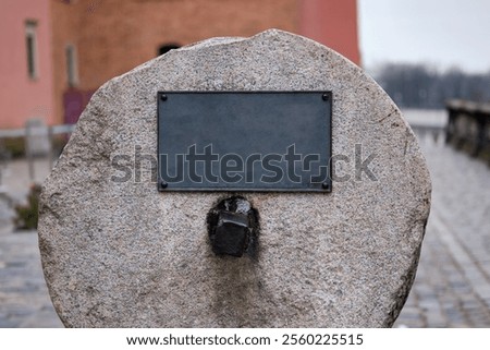 A large stone marker with an empty plaque is set in a riverside area, showcasing its historical significance amid a peaceful urban landscape. Overcast skies add to the tranquil atmosphere.