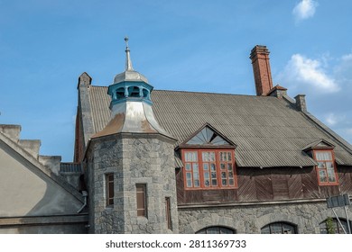 A large stone structure featuring a unique metal spire, a tall brick chimney, and dark wood paneling with bright red window frames under a clear blue sky.
