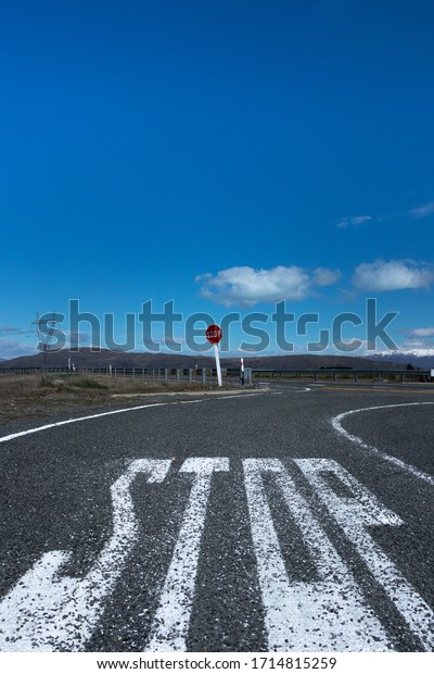 Large 'stop' lettering painted on tarmac
road & red sign post viewed from low level with blue sky,
clouds & snow capped mountains in
background