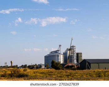 Large storage silos and processing structures stand in a field under a blue sky. The setting reflects an agricultural site with machinery and green fields surrounding the buildings.