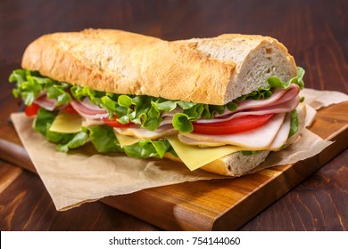 Large Subway Baguette Sandwich Cut In Half Filled With Ham, Turkey Breast, Cheese, Lettuce And Tomatoes On A Cutting Board Closeup.