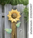 A large Sunflower made out of metal,complete with green leaves. The flower is mounted to the side of a light brown backyard fence.