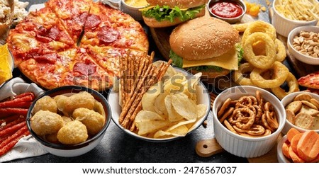 Large table of assorted take out food such as pizza, french fries, onion rings, burgers and snacks on a dark background. Top view.