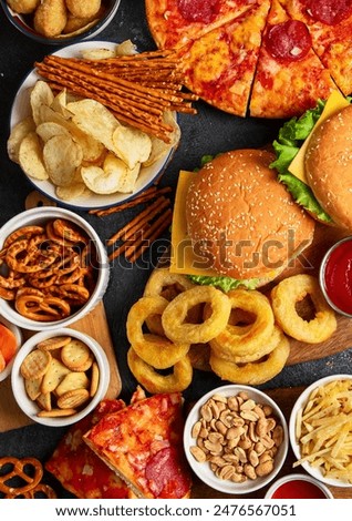 Large table of assorted take out food such as pizza, french fries, onion rings, burgers and snacks on a dark background. Top view.