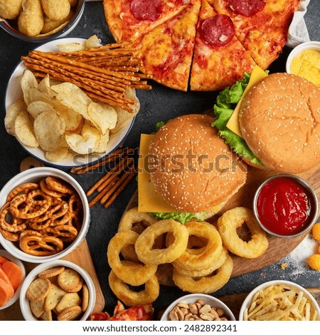 Large table of assorted take out food such as pizza, french fries, onion rings, burgers and snacks on a dark background. Top view.