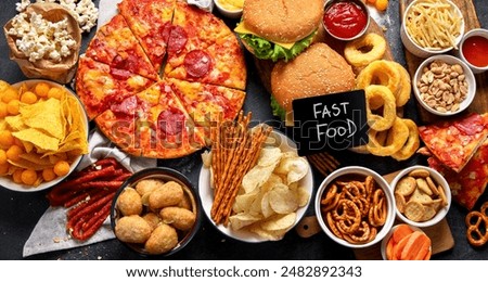 Large table of assorted take out food such as pizza, french fries, onion rings, burgers and snacks on a dark background. Top view.