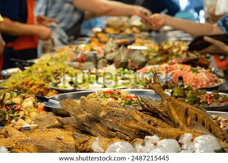 A large table with many different types of food, including fish. The table is crowded with people, some of whom are reaching for food. Scene is lively and social