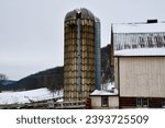 a large, tall silo with a pattern on it next to a large tan wooden barn with a brick base on a farm on a snowy day in the country 