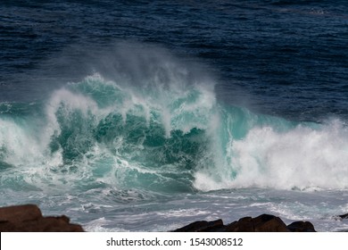 A Large Teal Green Colour Massive Rip Curl Wave As It Reaches The Rocky Shore. There's White Frothy Water With A Textured Water Spray Blowing Off The Tip Of The Wave With A Dark Blue Ocean.