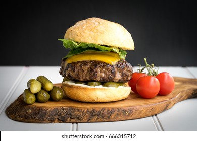 Large Thick Grilled Beef Minced Burger Patty With Toasted Bun Melted Cheddar Cheese Green Fresh Salad Cherry Tomato And Pickle Cucumber On Rustic Wooden Board And White Table