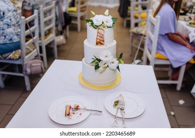 A Large Three-tiered White Cake With A Cut Off Piece On A Plate With A Fork And Knife Stands On A Table In A Restaurant. Food Photography, Wedding.