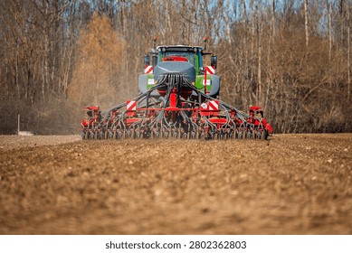 A large tractor pulls a wide pneumatic seed drill with hoses and coulters, forming even rows in a tilled field, bare deciduous trees beyond, daylight, low angle