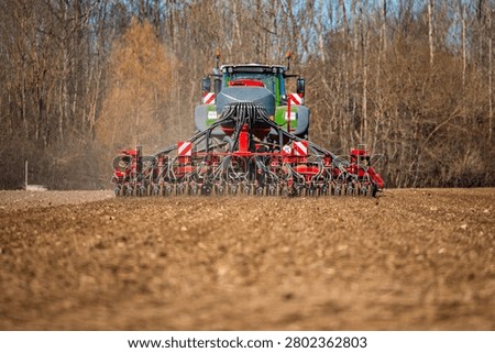 A large tractor pulls a wide pneumatic seed drill with hoses and coulters, forming even rows in a tilled field, bare deciduous trees beyond, daylight, low angle