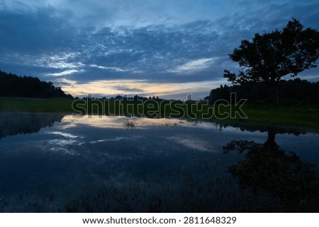 A large tree silhouette reflected in a calm rural pond under dramatic pre-dawn clouds and warm golden tones on the horizon in the Selija region of Latvia.