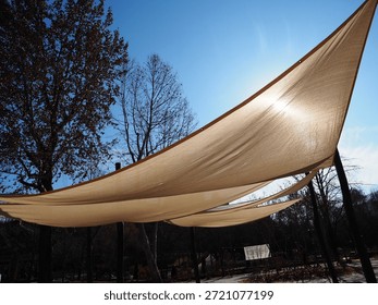 Large triangular sunshades filter bright sunlight in a park under a clear blue sky with tall trees.