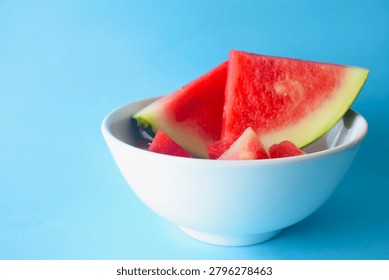 Large triangular watermelon slices and smaller chunks creatively arranged inside a white ceramic bowl against a blue background.
