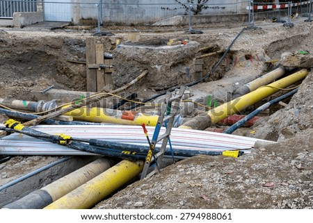 Large utility pipes, cable conduits, and a ladder span an open trench at a road construction site with exposed soil and infrastructure work underway.