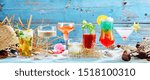 Large variety of tropical cocktails, martini and whisky in a panorama banner arranged on white sea sand over a rustic blue wood background with copy space