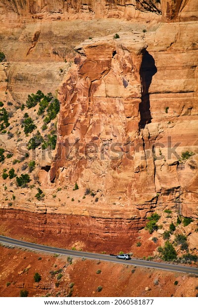 Large vertical red wall of rock with tiny road\
and car cutting through