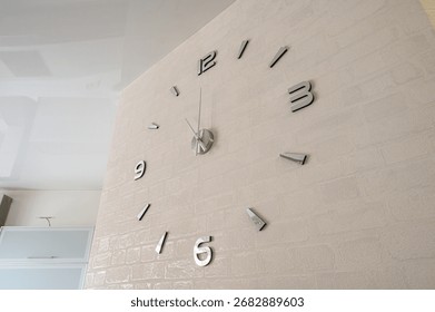 large wall clock with silver numbers on a beige, textured wall. The clock face is minimalist