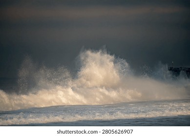 A large wave explodes into a spray of water with a pier visible in the distance on a dark, overcast day