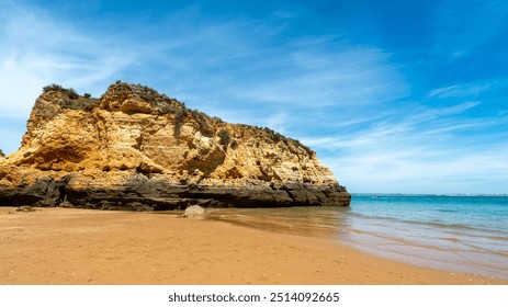 A large, weathered rock formation towers over a sandy beach in Algarve, Portugal. The golden hues of the cliff face contrast against the blue sky and turquoise waters.  - Powered by Shutterstock