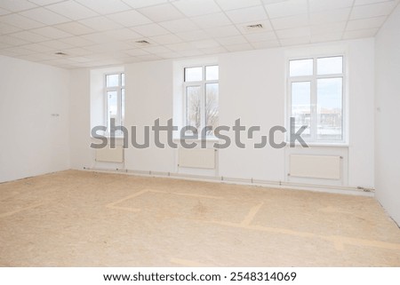 A large, well-lit empty room with white walls, ample natural light from windows, and ceiling lights. Ideal for renovation or office setup concepts.
