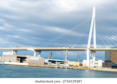 Large white cable-stayed bridge spanning over a harbor with industrial port buildings and cranes in the background. Modern infrastructure and maritime transport scene under a cloudy sky.