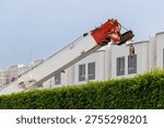 Large white crane with red hook lifts heavy load above green hedge. Modern building under construction with visible scaffolding. Scene shows active industrial development. Image ideal for construction