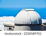 A large white dome on top of a building. The dome is surrounded by clouds and the sky is blue