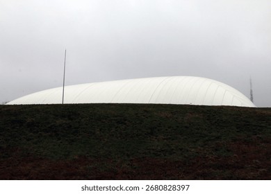 Large white dome structure behind a grassy hill under an overcast sky, with a tall pole and distant communication tower, evoking modern architecture and landscape contrast.