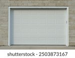 A large white metal car garage door on a grey brick house. The solid single steel door has square patterns or panels. The modern luxury automotive building has a grey concrete driveway entrance. 