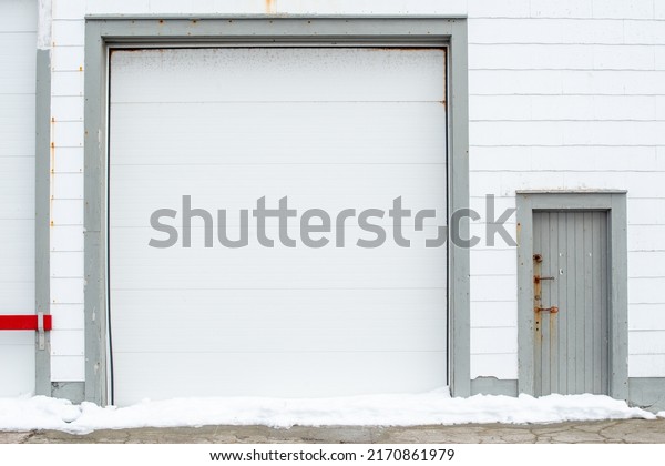 A large white metal panel car garage door with dark
gray trim and white clapboard siding. A single wooden door and
entrance to the building is to the right of the garage door with
multiple rusty locks