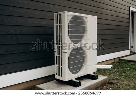 A large white outdoor air conditioner stands on a betaine base against a black wooden wall until it is hung in the air conditioner space.