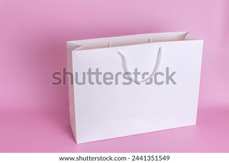 A large white paper bag with white rope handles stands on a pink background in close-up.